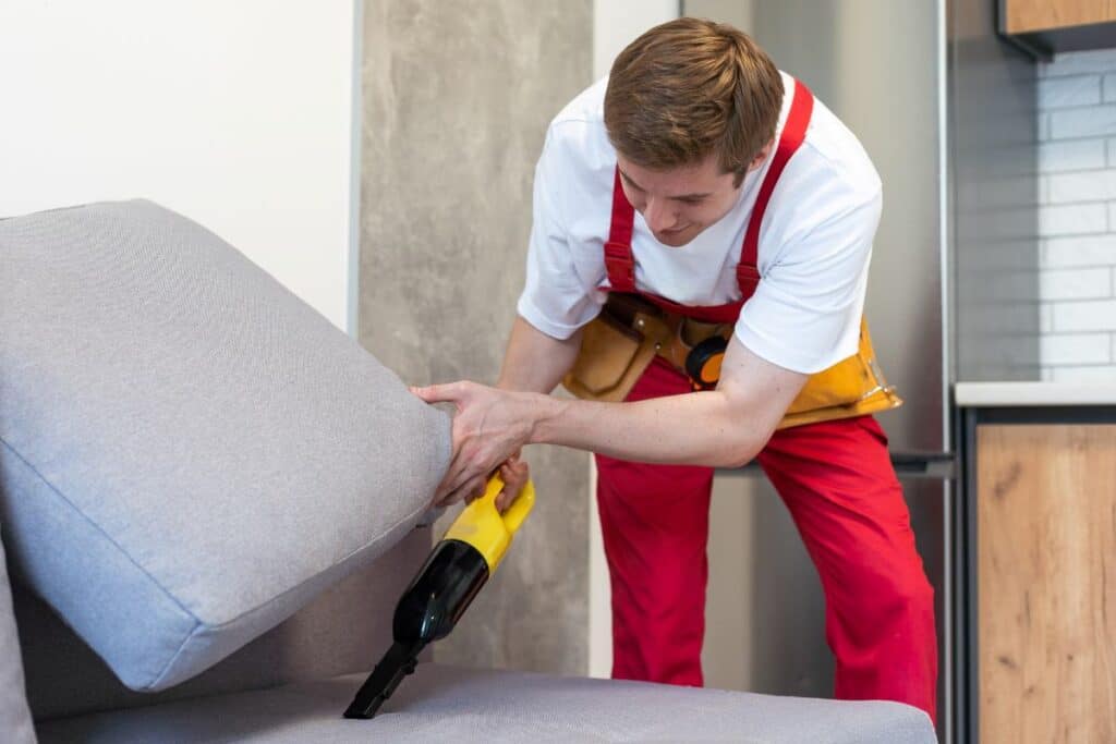 Rear View Of Young Male Worker Cleaning Sofa With Vacuum